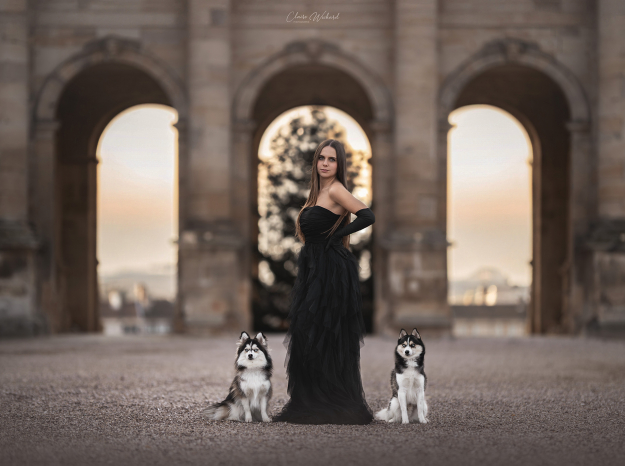 Portrait d'une femme en robe de soirée noire avec ses deux chiens Pomsky devant les arcades du Château de Lunéville