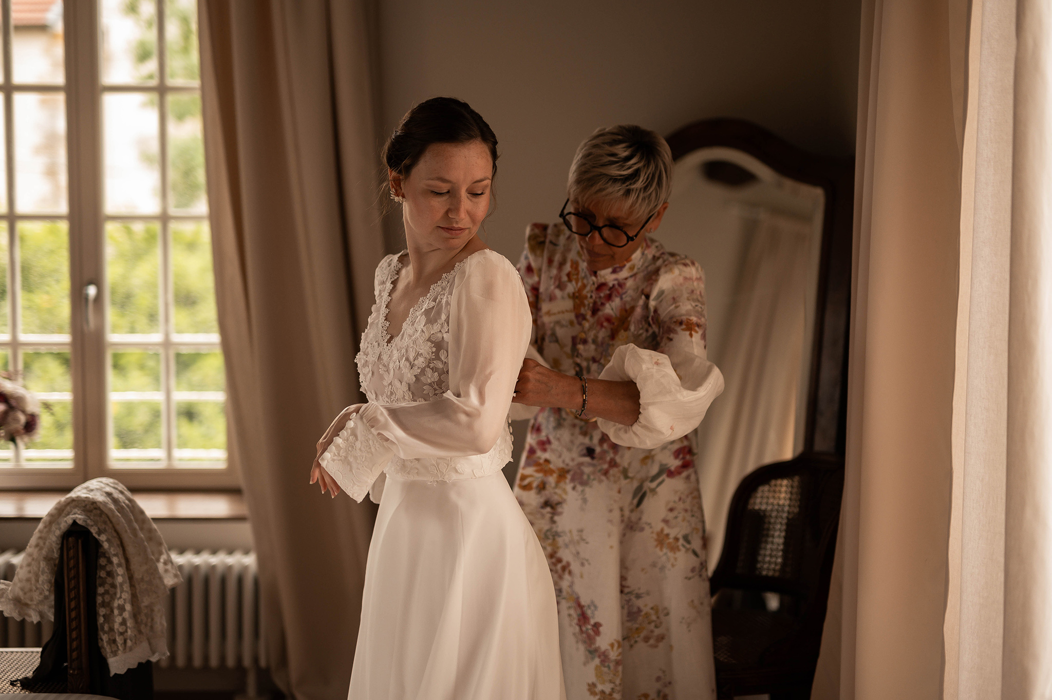 La maman de la mariée aidant sa fille à boutonner sa robe de mariée en dentelle au Château Ferme d'Aboncourt