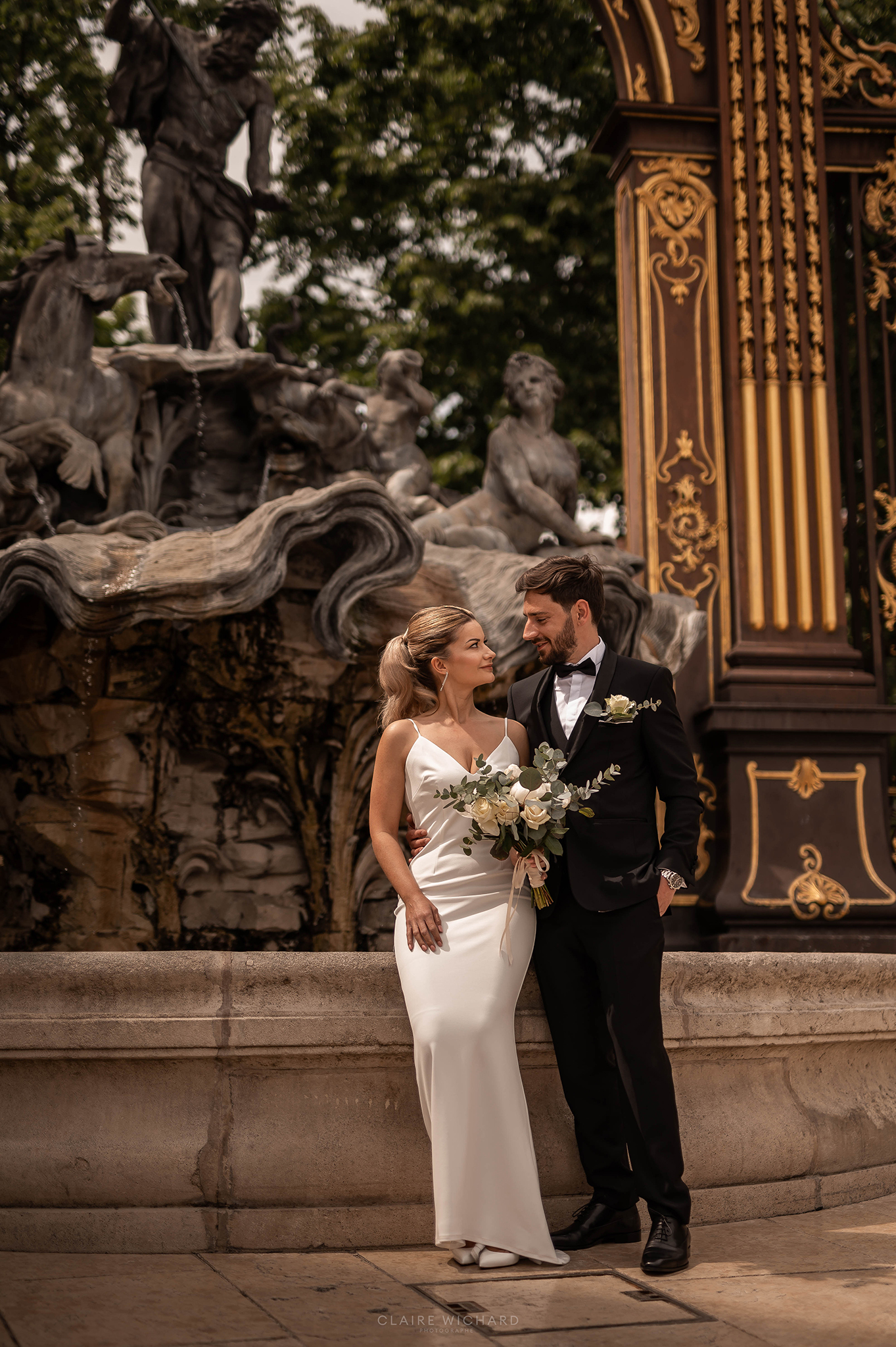 Mariés posant devant la fontaine d'Amphitrite et les grilles dorées de la Place Stanislas à Nancy sous l'œil de leur photographe claire wichard