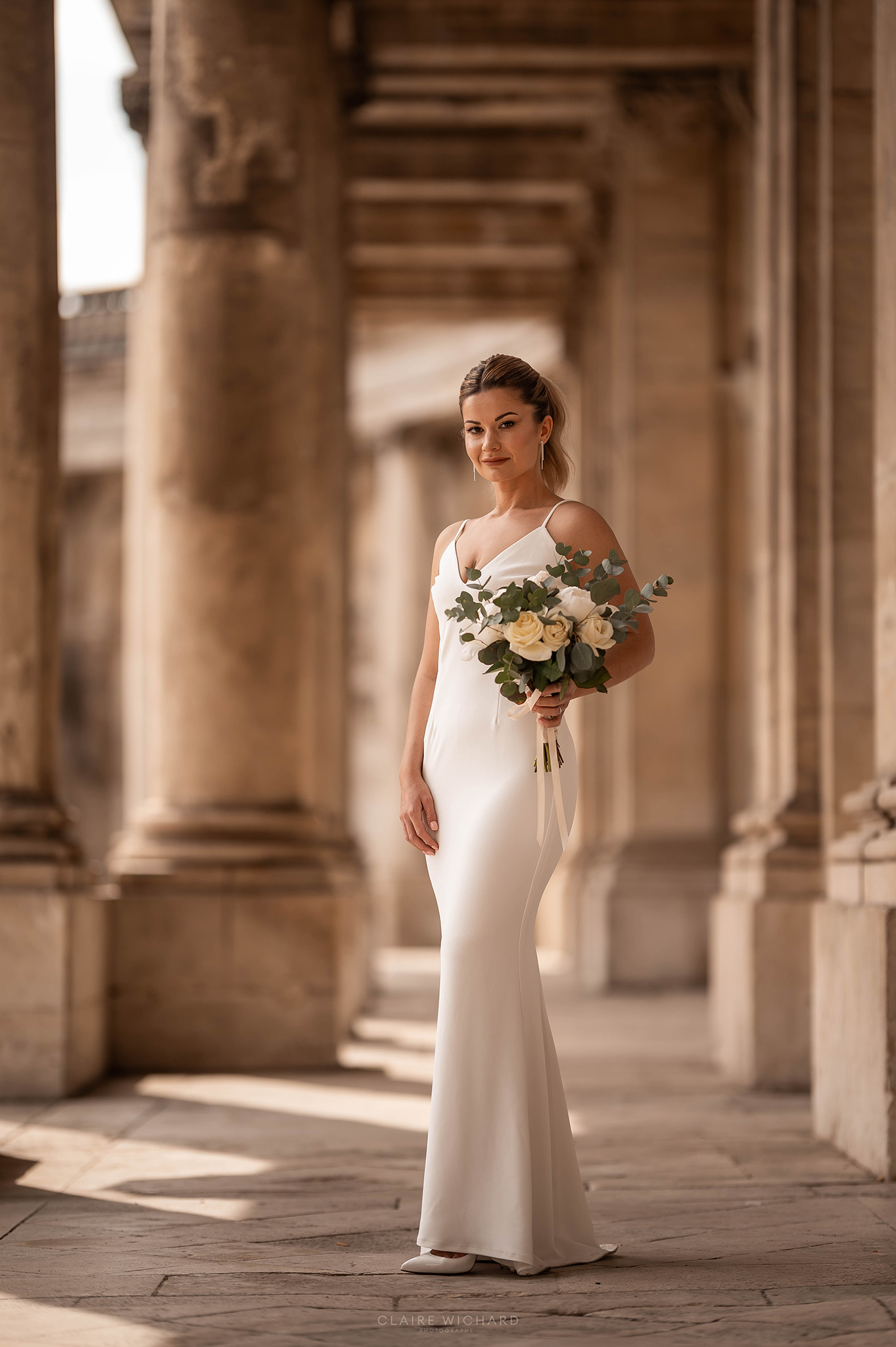 Portrait en pied d'une mariée avec son bouquet sous les colonnades historiques du Palais du Gouvernement à Nancy