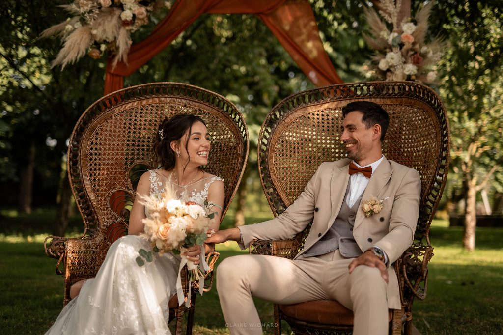 Couple de mariés souriant assis dans des fauteuils en osier lors d'une cérémonie laïque en extérieur au Château Ferme d'Aboncourt