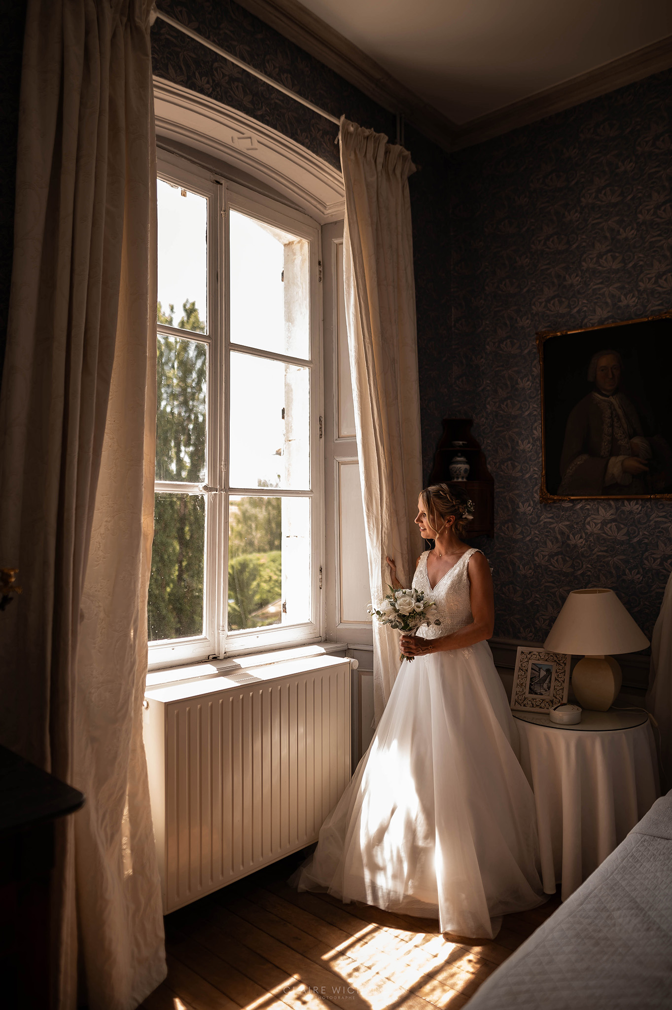 La mariée en robe blanche devant une grande fenêtre d'époque à l'intérieur du Château d'Autigny-la-Tour.