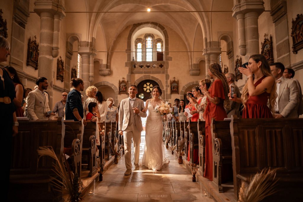 La mariée et son papa remontant l'allée centrale de l'église d'Aboncourt, sous les applaudissements des invités, style reportage mariage Nancy