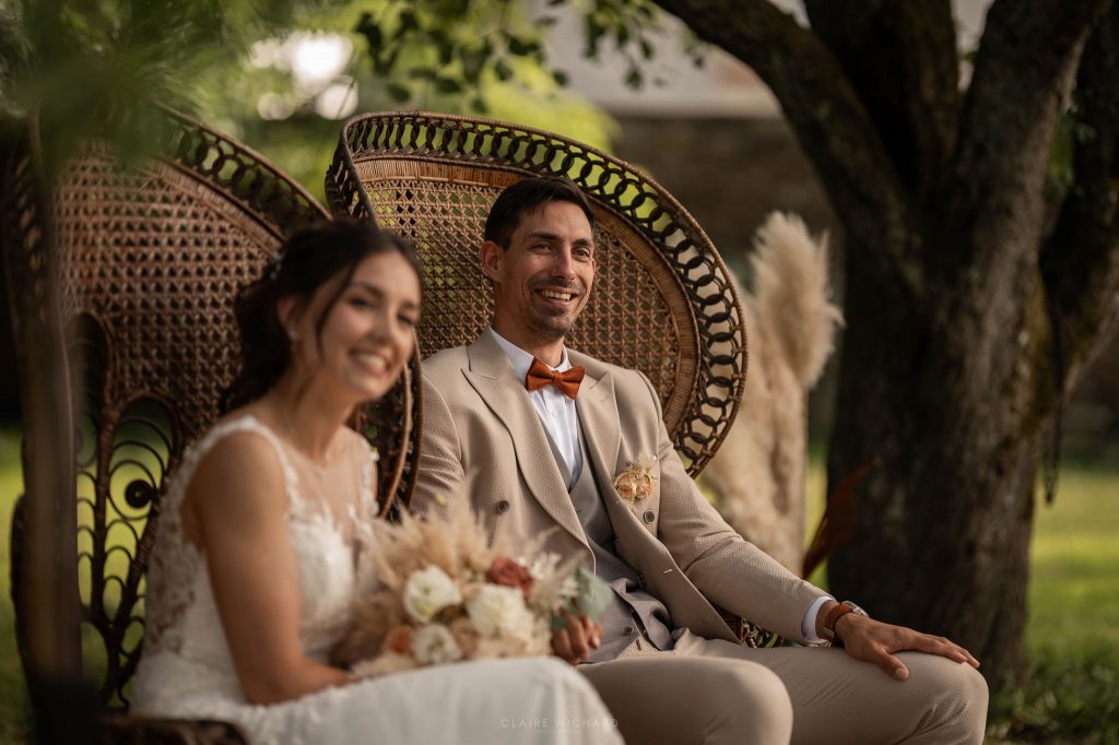 Portrait des mariés souriants installés dans des fauteuils en rotin (Emmanuelle) pendant la cérémonie laïque, lors d'un mariage bohème chic au Château ferme d'Aboncourt