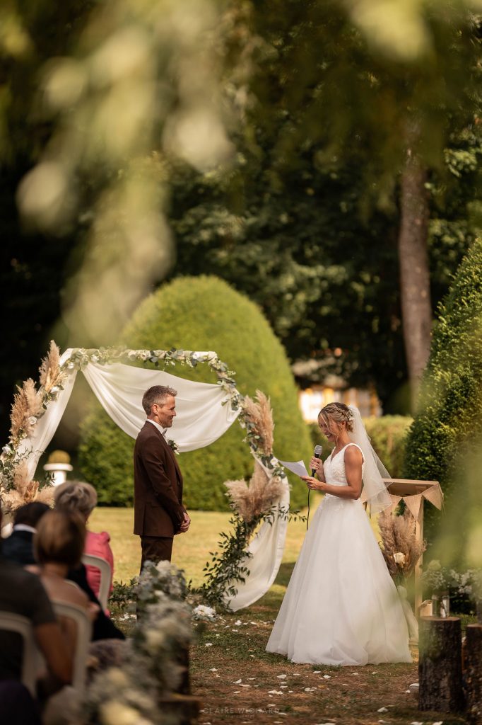 Cérémonie laïque dans les jardins du Château d'Autigny-la-Tour : la mariée lit ses vœux au micro sous une arche fleurie, photographiée par Claire Wichard.