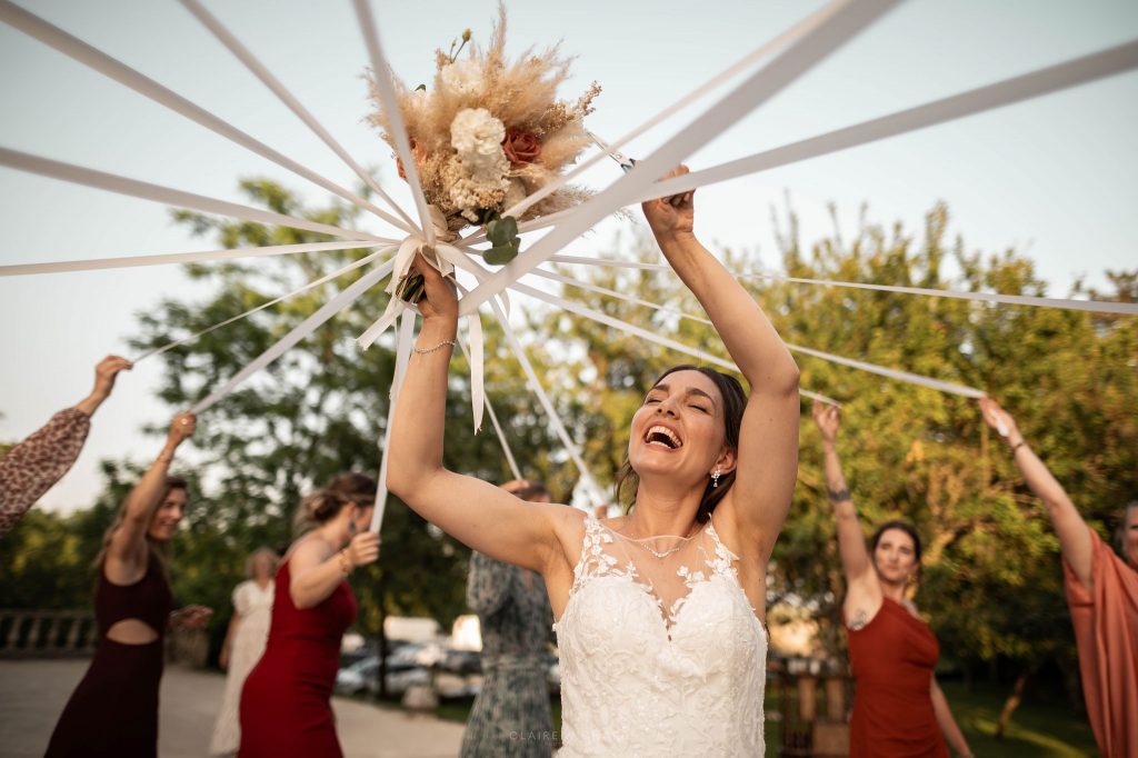 Moment de joie capturé au château ferme d'Aboncourt près de Nancy par Claire Wichard : la mariée en robe de dentelle lève le bouquet de fleurs séchées pendant le jeu des rubans avec ses invitées.