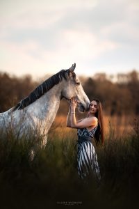 Détail d'une femme caressant le museau d'un cheval gris pommelé en lumière de fin de journée.