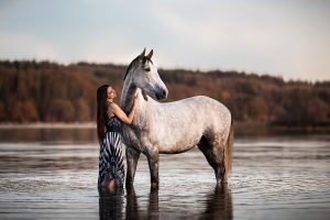 Femme en robe longue debout dans l'eau avec son cheval gris, reflets au coucher du soleil à Bouzey.