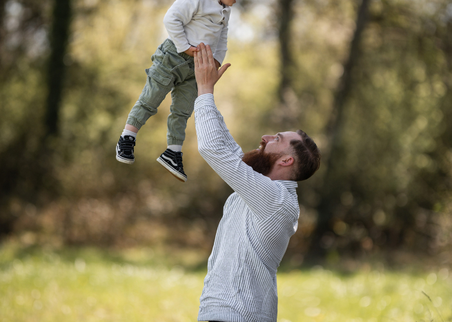 Un père joyeux lance son jeune enfant en l'air dans un bois baigné de lumière naturelle. Photographie de famille spontanée à Nancy par Claire Wichard.