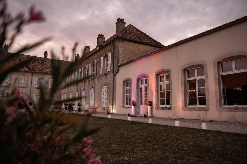 Photographie de Claire Wichard de la façade illuminée en rose et blanc du Château d'Autigny-la-Tour, avec des fleurs en premier plan, capturant l'ambiance d'une soirée de mariage élégante près de Nancy.