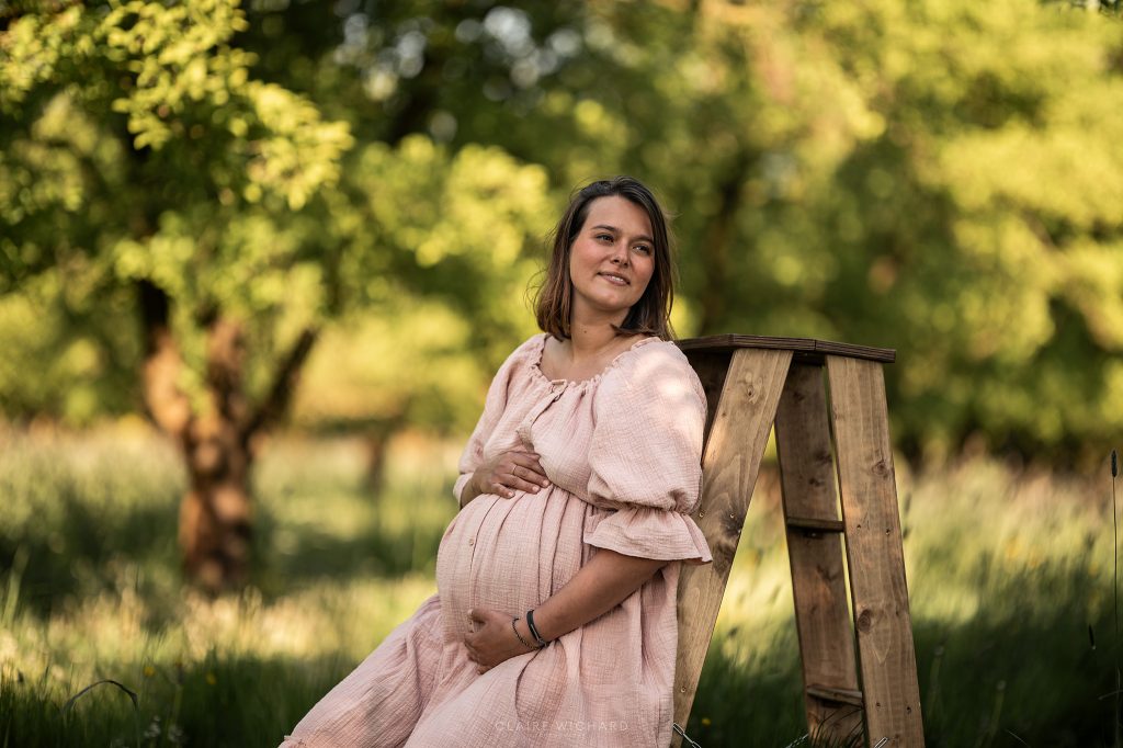 Femme enceinte en robe rose poudré posant dans un verger de mirabelliers près de Nancy pour une séance photo grossesse lumineuse en extérieur.