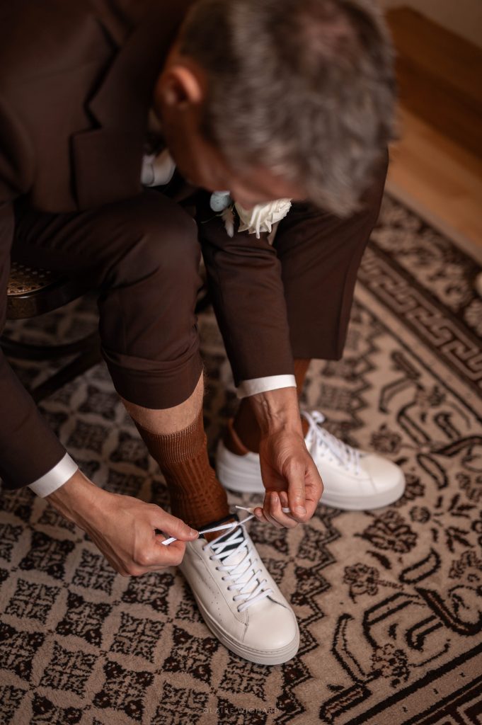 Le marié en costume marron mettant ses baskets blanches, préparatifs chic et décontractés au domaine