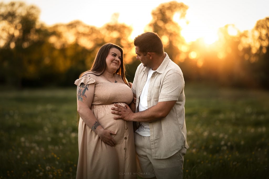 Séance photo couple et maternité dans un style chaleureux. La femme enceinte regarde son compagnon avec tendresse tandis qu'il caresse son ventre rond au milieu d'un champ en Meurthe-et-Moselle