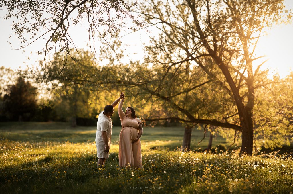 Séance photo de grossesse en extérieur près de Nancy lors du coucher de soleil. Un futur papa fait tourner délicatement la future maman enceinte dans une prairie en fleurs sous une lumière dorée et chaleureuse