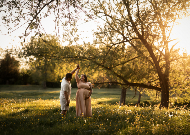 Séance photo de grossesse en extérieur près de Nancy lors du coucher de soleil. Un futur papa fait tourner délicatement la future maman enceinte dans une prairie en fleurs sous une lumière dorée et chaleureuse