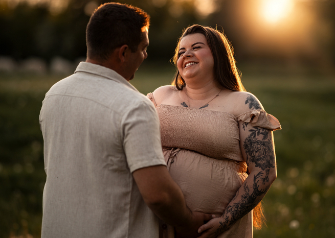 Gros plan lumineux d'une future maman souriante et tatouée lors d'une séance photo de grossesse à Nancy. Le futur papa pose tendrement ses mains sur son ventre arrondi sous la lumière dorée du soleil couchant.