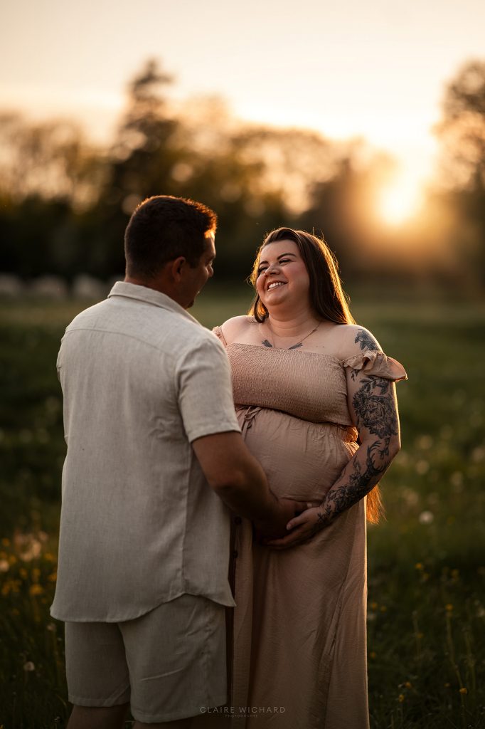 Gros plan lumineux d'une future maman souriante et tatouée lors d'une séance photo de grossesse à Nancy. Le futur papa pose tendrement ses mains sur son ventre arrondi sous la lumière dorée du soleil couchant.