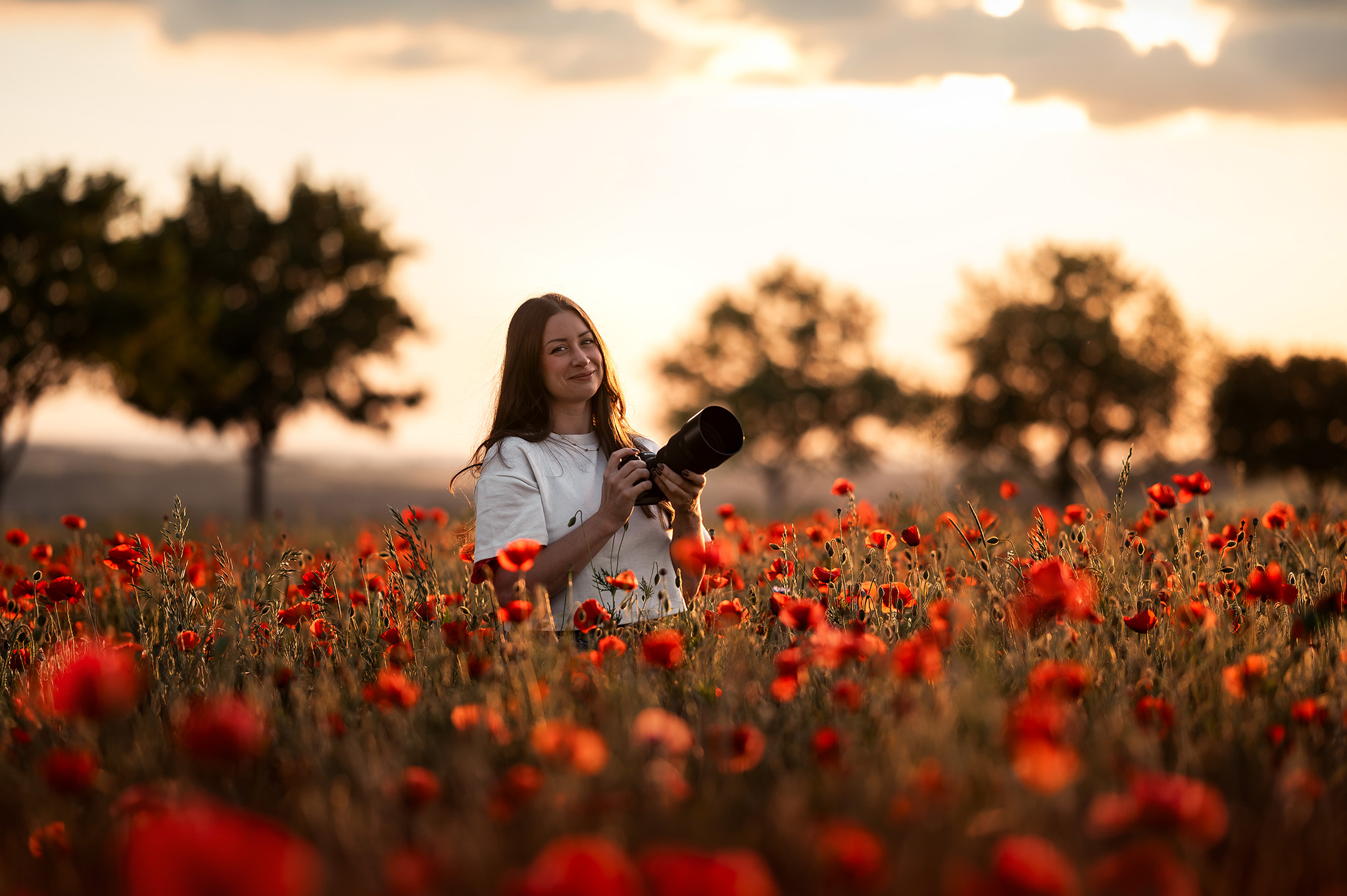 Portrait de Claire Wichard, photographe professionnelle souriante, son appareil photo à la main, debout au milieu d'un grand champ de coquelicots rouges en Lorraine lors d'un coucher de soleil lumineux.