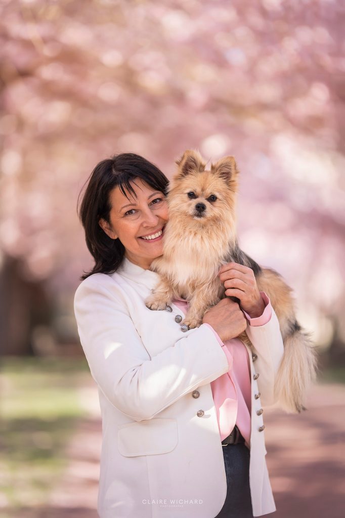Portrait complice d'une femme brune câlinant son petit chien beige sous les fleurs roses.