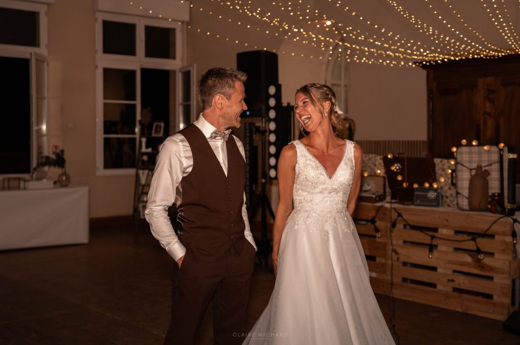 Photo de Claire Wichard montrant un moment de joie authentique : le marié en gilet marron et la mariée en robe en dentelle éclatent de rire ensemble sous des guirlandes lumineuses dans la salle de réception du Château d'Autigny-la-Tour.