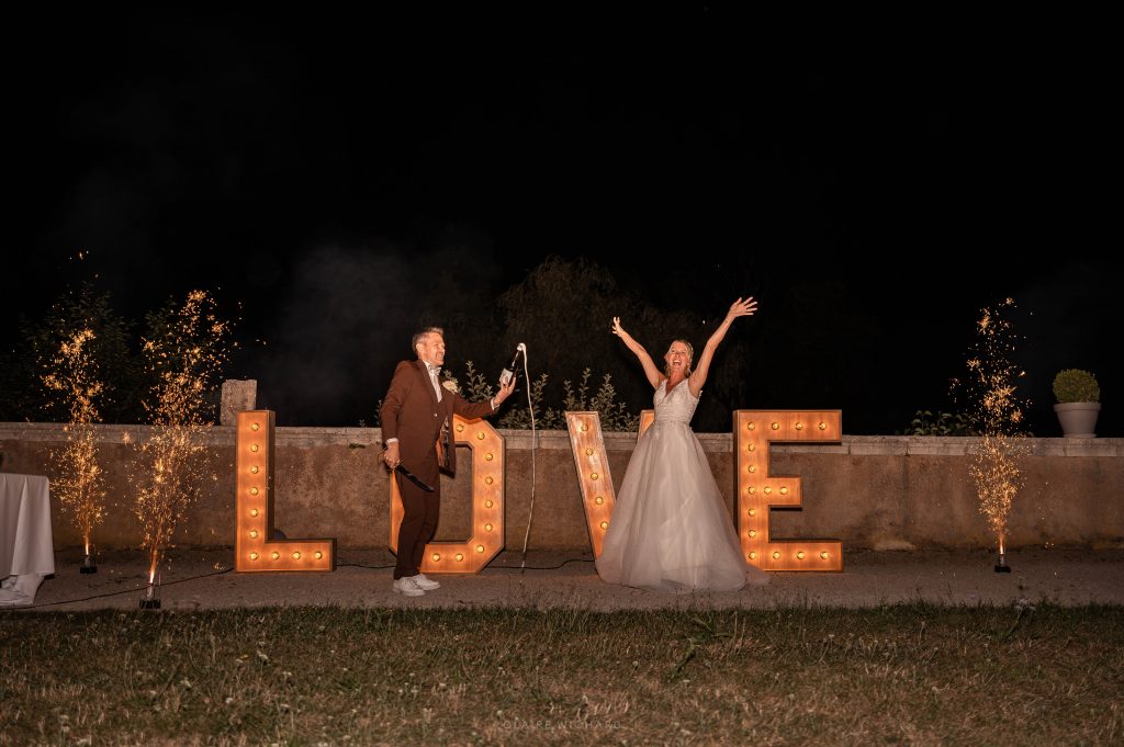 Photographie de mariage prise de nuit par Claire Wichard au Château d'Autigny-la-Tour. Le marié sabre une bouteille de champagne tandis que la mariée lève les bras en signe de célébration devant de grandes lettres "LOVE" lumineuses et des feux d'artifice de scène.