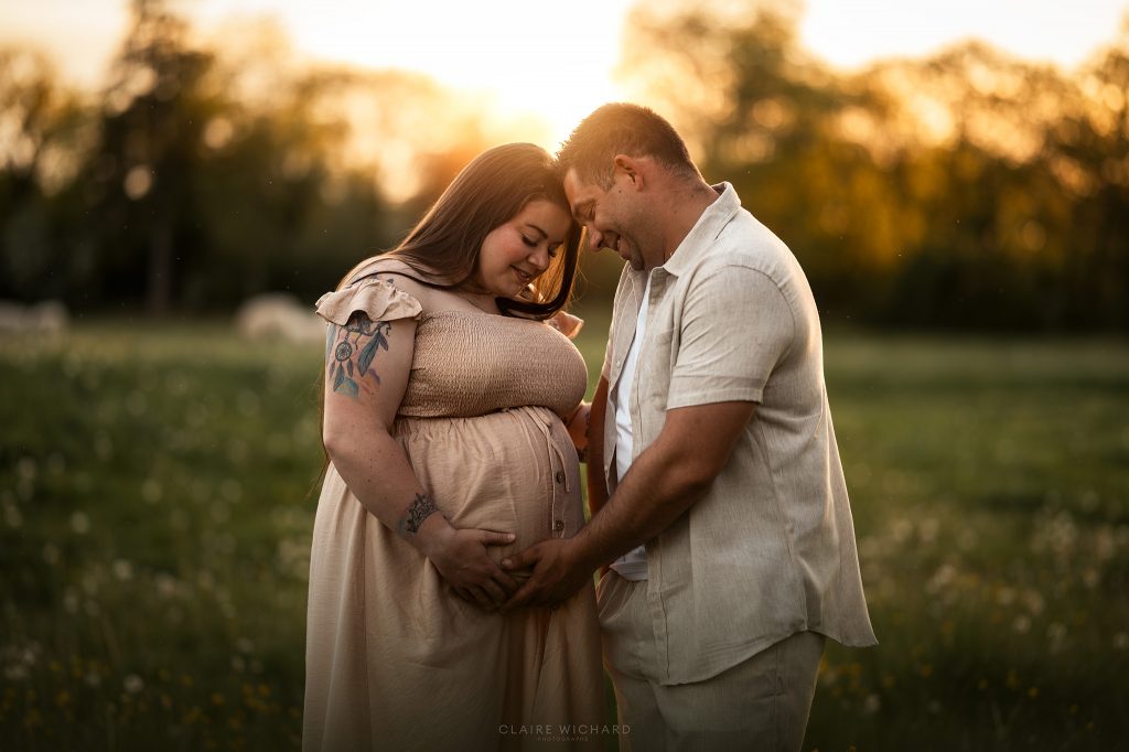 Moment de complicité et de douceur entre futurs parents lors d’un shooting grossesse en extérieur. Le couple pose front contre front, les mains réunies sur le ventre de la maman, avec un arrière-plan boisé et lumineux