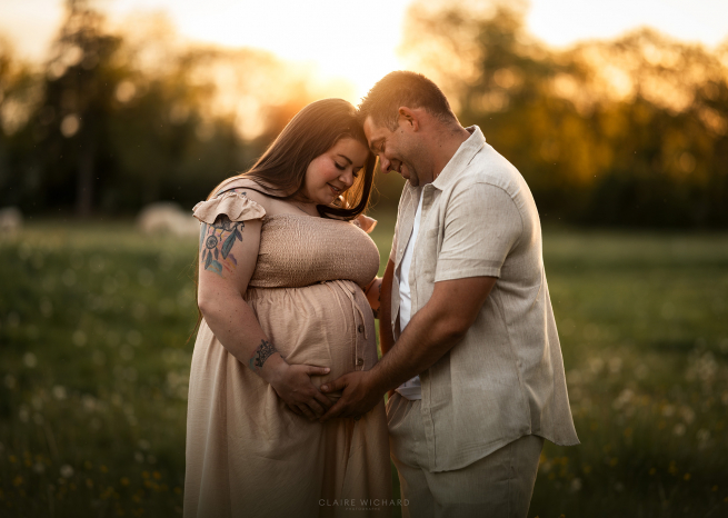 Moment de complicité et de douceur entre futurs parents lors d’un shooting grossesse en extérieur. Le couple pose front contre front, les mains réunies sur le ventre de la maman, avec un arrière-plan boisé et lumineux