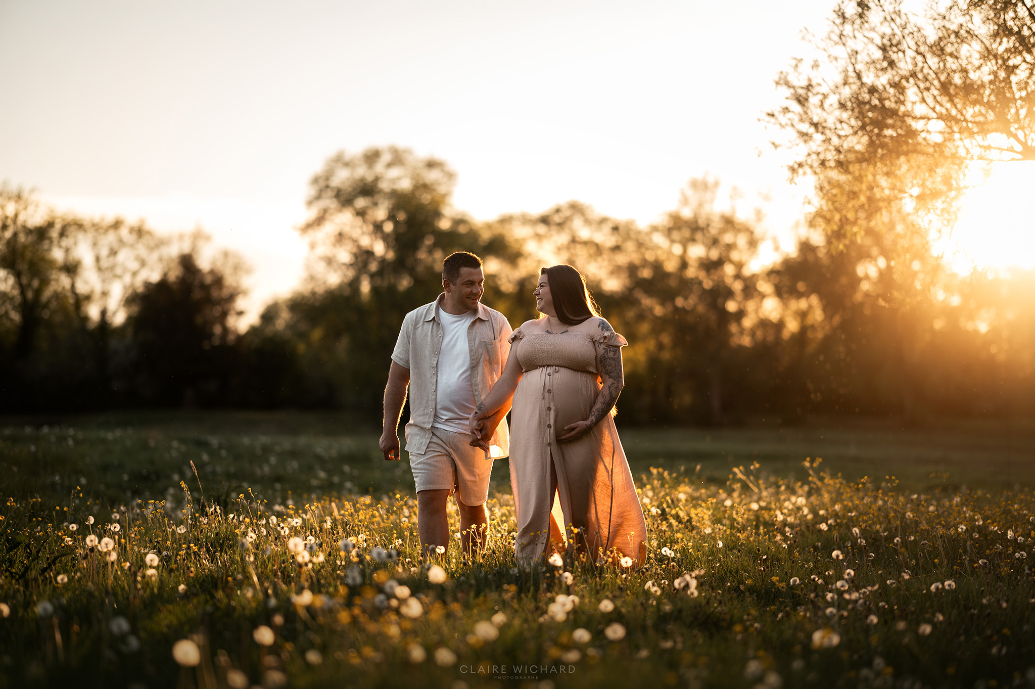 Portrait de couple pendant une séance de grossesse en pleine nature aux alentours de Nancy. Le futur père et la femme enceinte en robe bohème beige se regardent avec amour en marchant au milieu des fleurs sauvages.