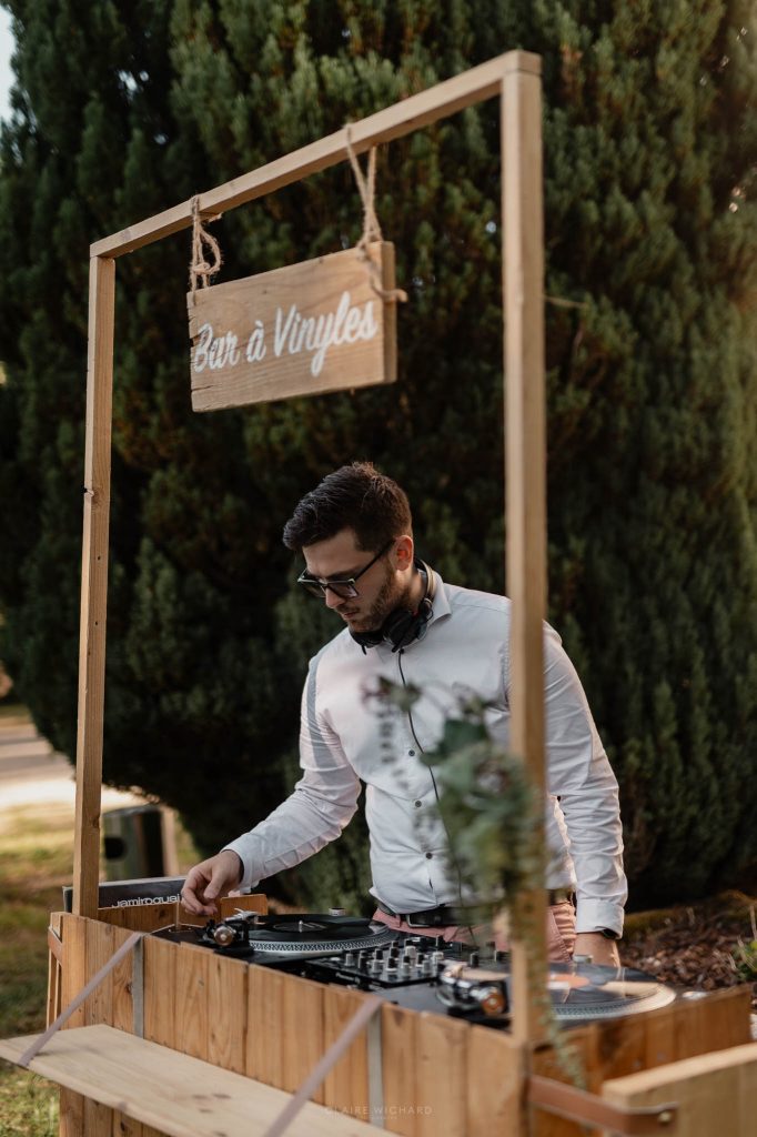 Un DJ de mariage mixe derrière un bar à vinyles en bois installé en extérieur lors d'une réception.