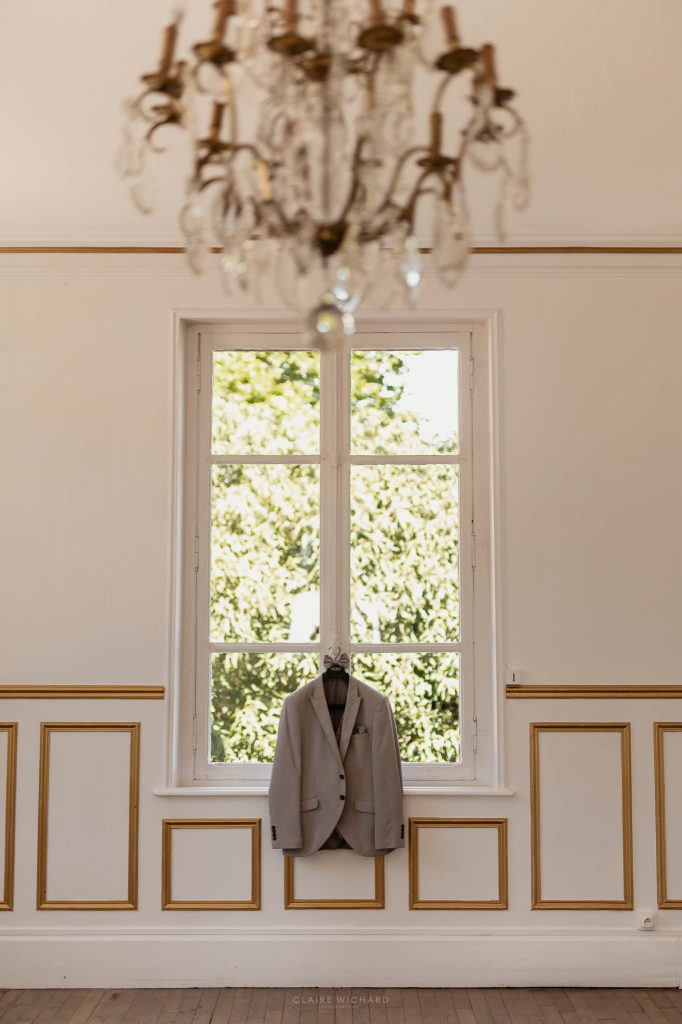 Costume de marié beige suspendu à une fenêtre lumineuse avec moulures dorées au Château des Aulnes