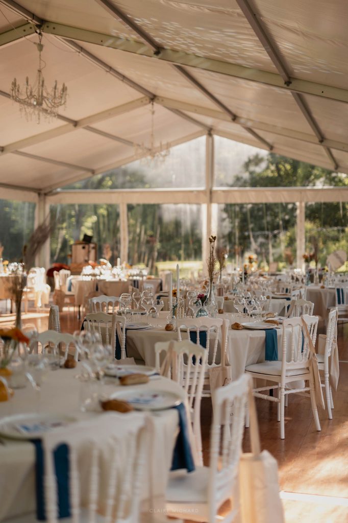 Tables de mariage décorées avec des nappes blanches, des chaises en bois blanc et des touches de bleu pétrole sous un chapiteau ensoleillé au chateau des aulnes