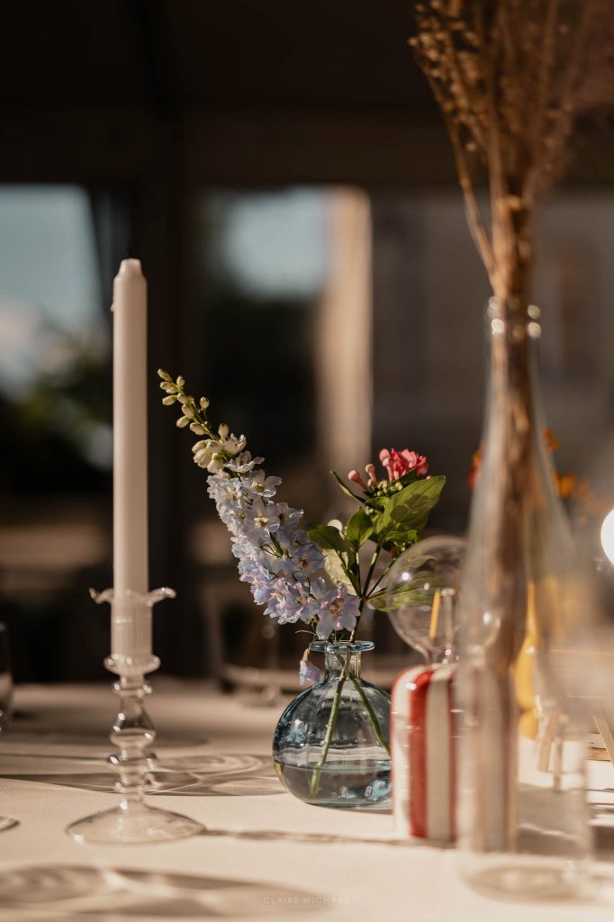 Détail de décoration de table de mariage avec un petit vase bleu contenant des fleurs de delphinium et une bougie blanche au château des aulnes