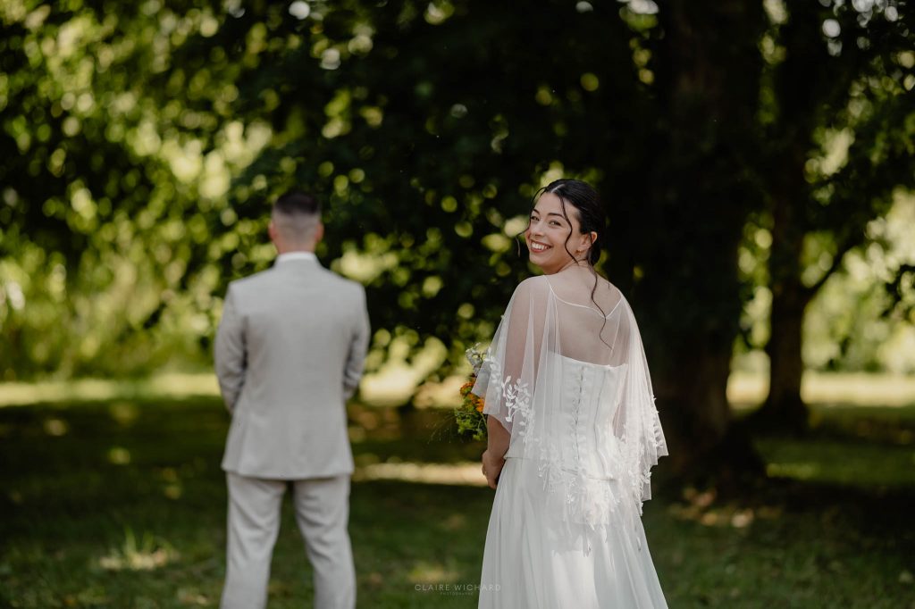 La mariée se retourne en souriant vers le marié de dos lors de leur séance first look dans le grand parc du château des aulnes