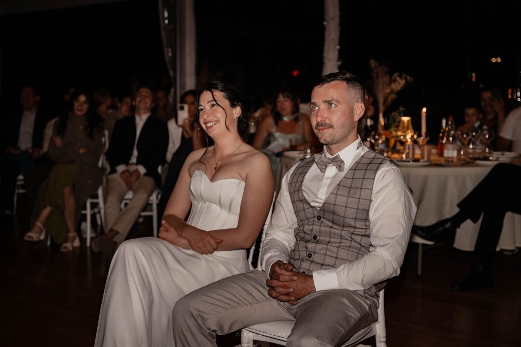 Les mariés assis à leur table d'honneur écoutent avec émotion et sourire les discours de leurs proches pendant le dîner au chateau des aulnes