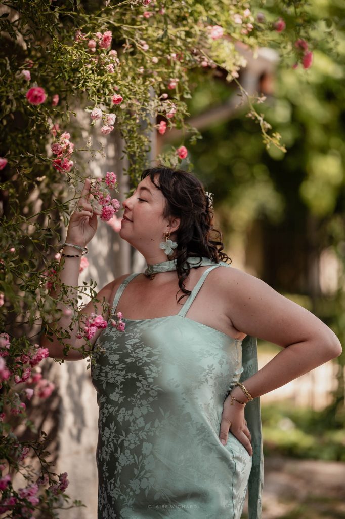 Portrait de profil d'une invitée en robe de satin vert pastel sentant des roses en fleurs près d'un mur en pierre à Nancy