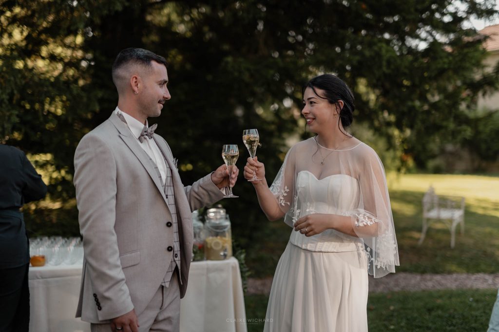Les mariés souriants trinquent avec une coupe de champagne à la main dans le parc pendant leur cocktail de mariage.