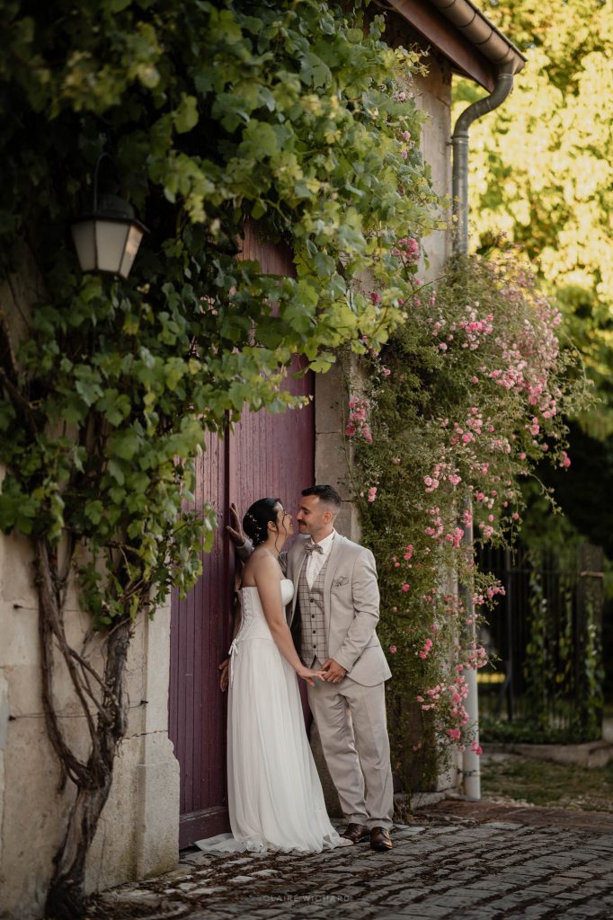 Les mariés se rapprochent pour un baiser contre une grande porte en bois bordeaux encadrée de rosiers grimpants en fleurs à Nancy