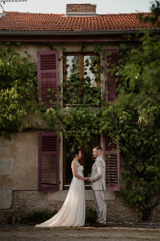 Les mariés se tiennent par la main devant la façade d'une maison ancienne couverte de vigne vierge aux volets pourpres