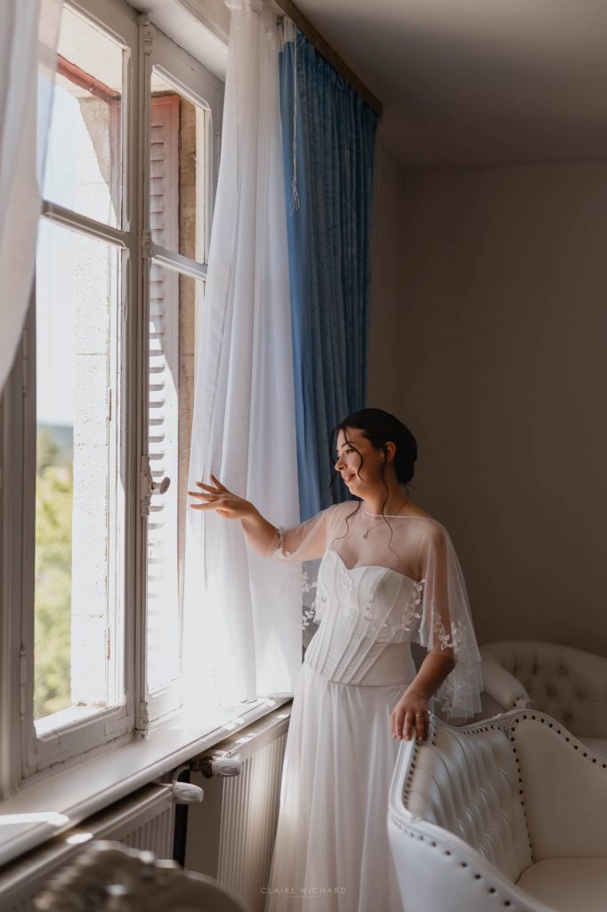 Portrait lumineux d'une mariée pensive regardant par la fenêtre du Château des Aulnes près de Nancy.