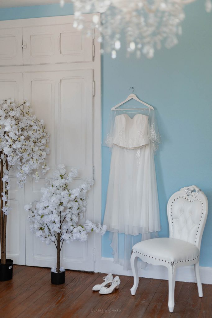 Robe de mariée blanche à dentelle suspendue dans une chambre romantique aux murs bleus au Château des Aulnes.