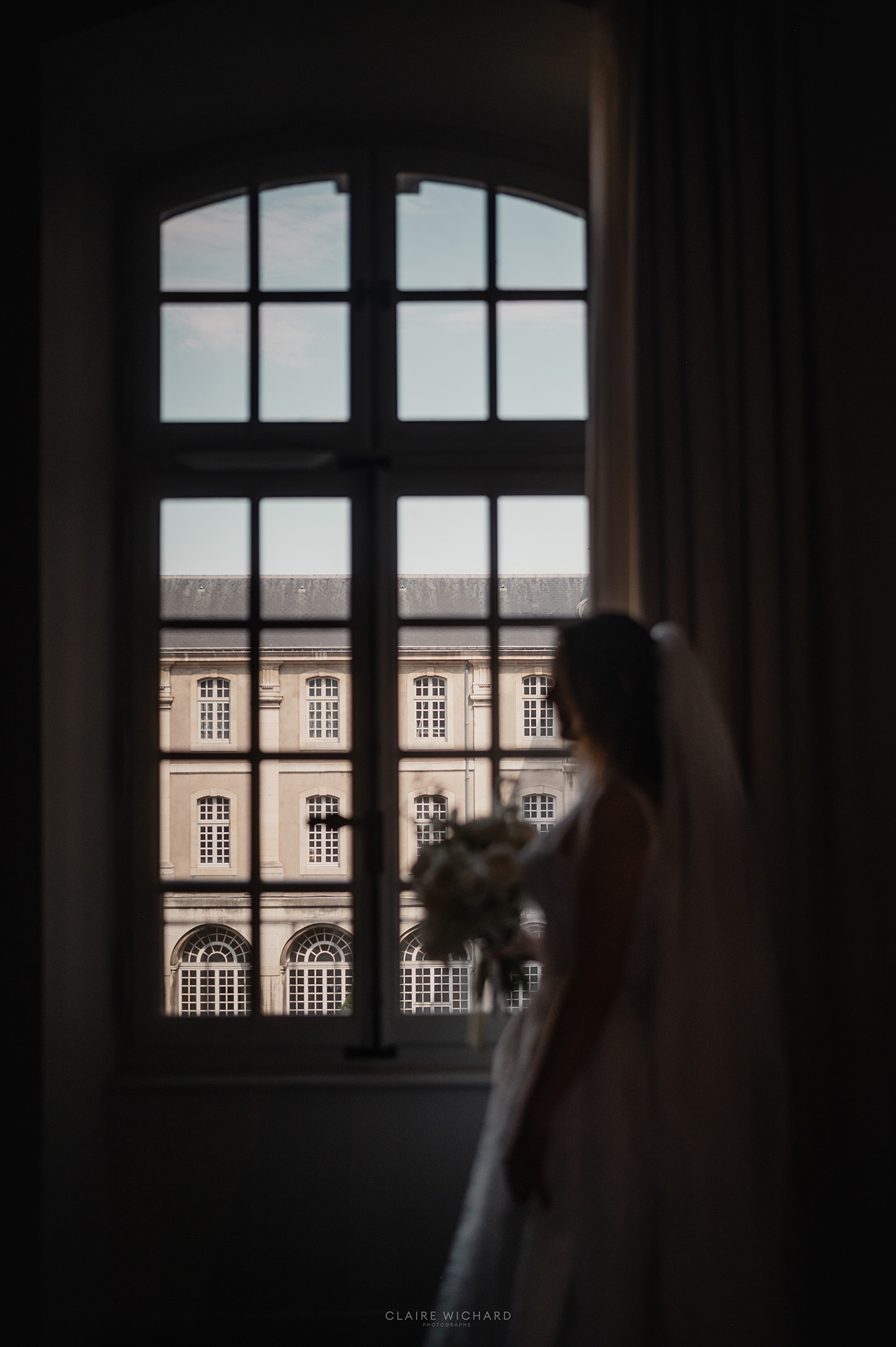 Silhouette de la mariée de profil tenant son bouquet, regardant par une grande fenêtre de l'Abbaye des Prémontrés près de Nancy, photo de Claire Wichard photographe mariage Lorraine
