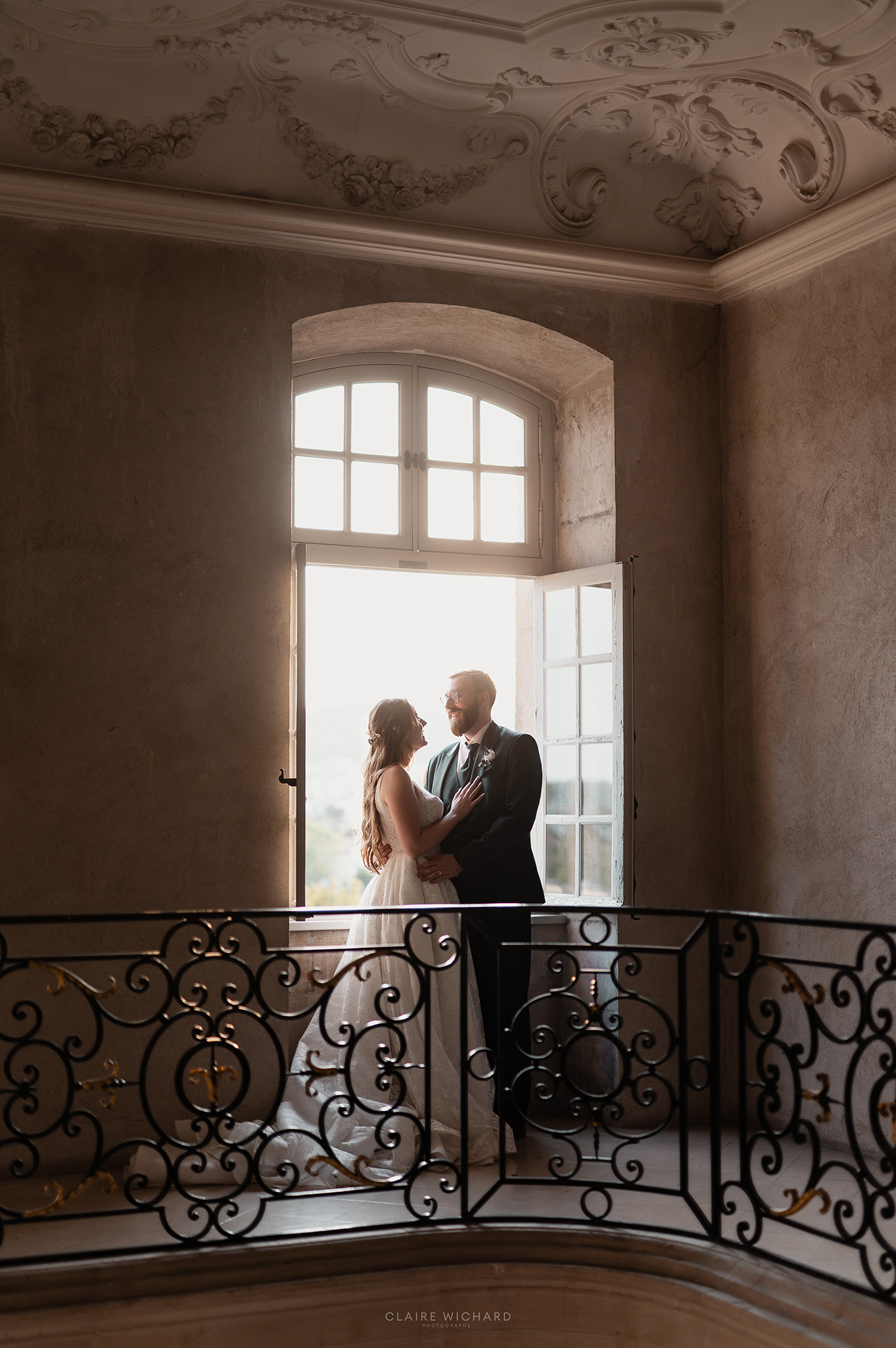 Le couple de mariés enlacé près d'une grande fenêtre lumineuse et d'une rampe en fer forgé à l'Abbaye des Prémontrés, reportage par Claire Wichard photographe Nancy