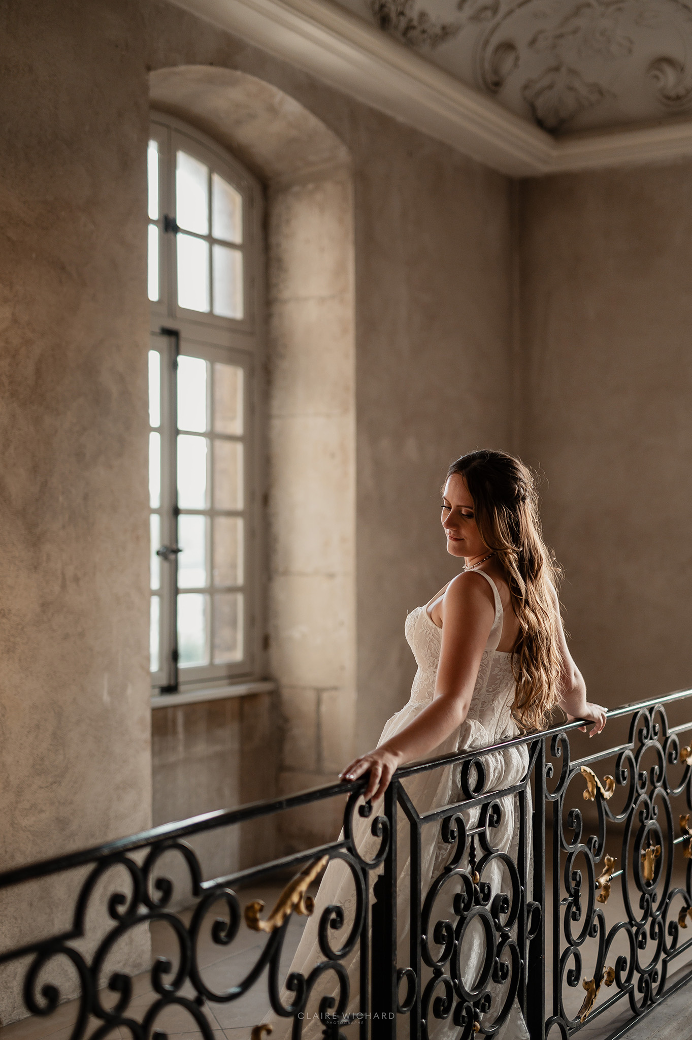 Portrait de la mariée souriante accoudée à la balustrade en fer forgé d'un étage historique de l'Abbaye des Prémontrés, séance photo de mariage par Claire Wichard en Lorraine